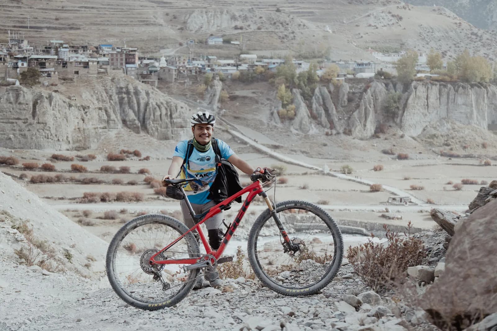 A smiling rider stands with a red bike above eroded cliffs, a stone village on the terraces behind.