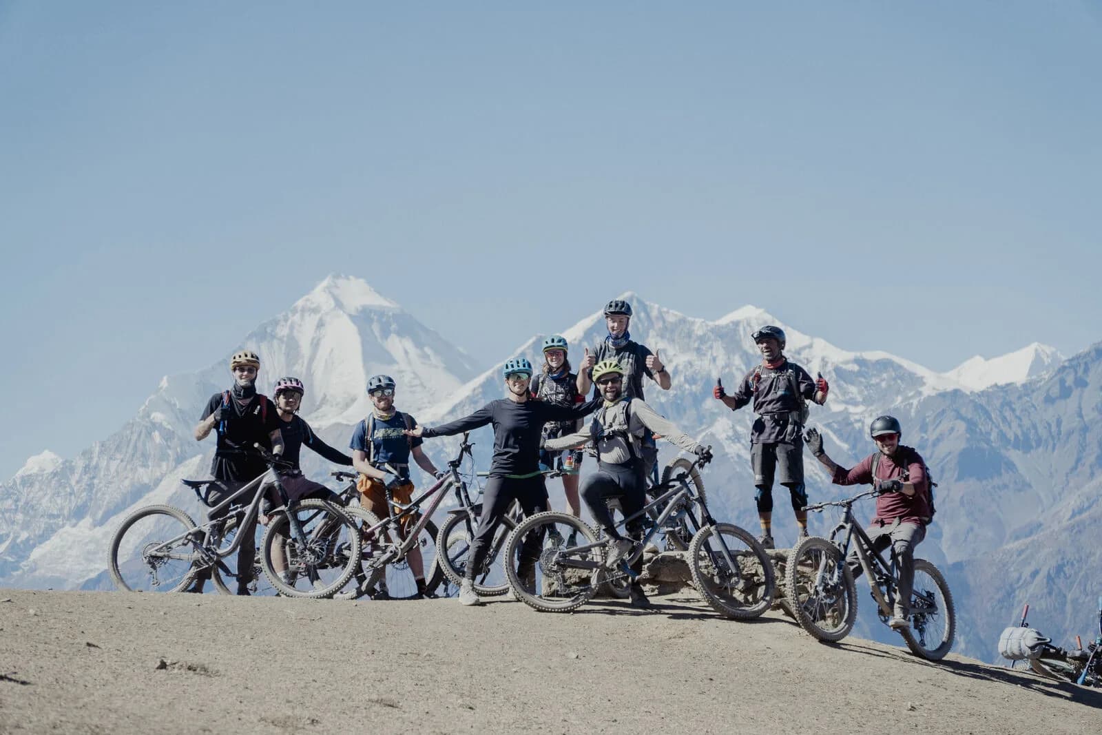 A group of riders lines a sandy crest with bikes raised, huge snow peaks on the horizon.