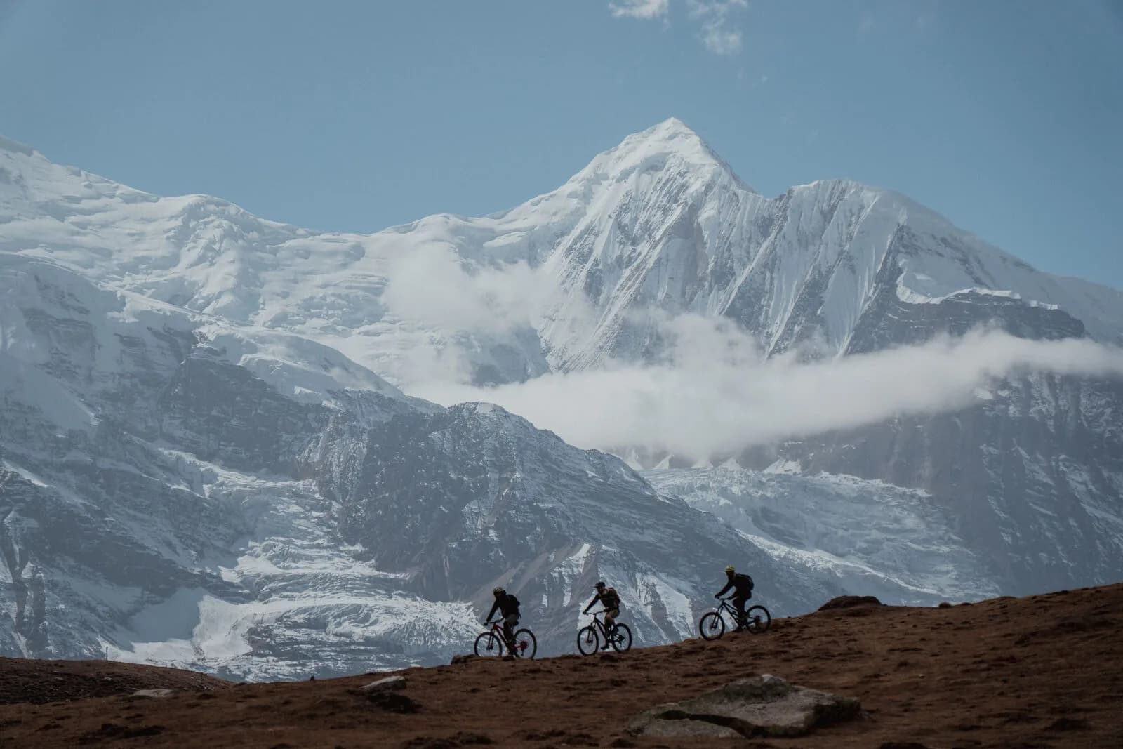 Three mountain bikers ride along a high brown ridgeline, silhouetted against a vast glaciated peak wrapped in cloud.