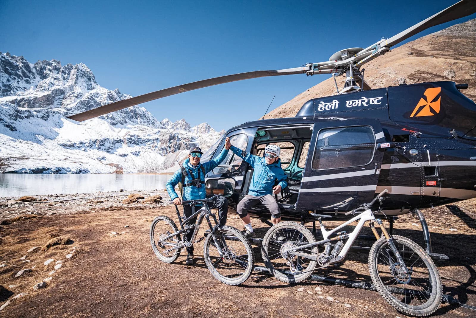 Two riders with their bikes beside a helicopter on brown ground by a high lake, snow peaks behind.