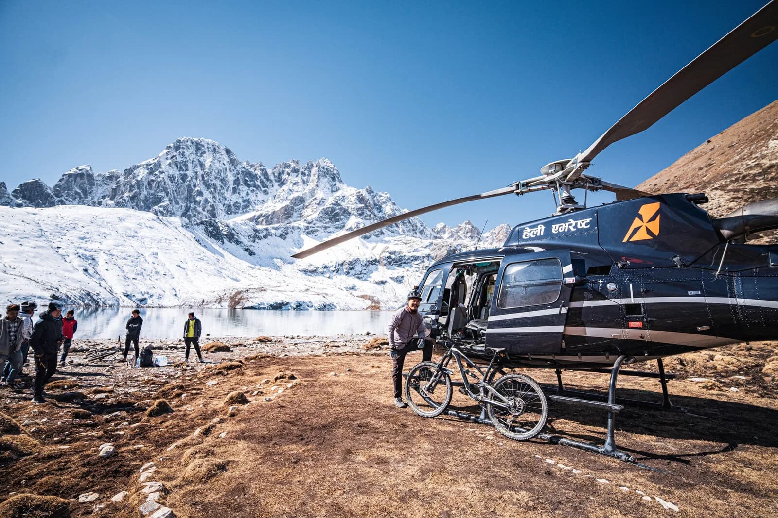 A helicopter stands on brown ground by a high lake below snow peaks, a bike unloaded beside it and people waiting.