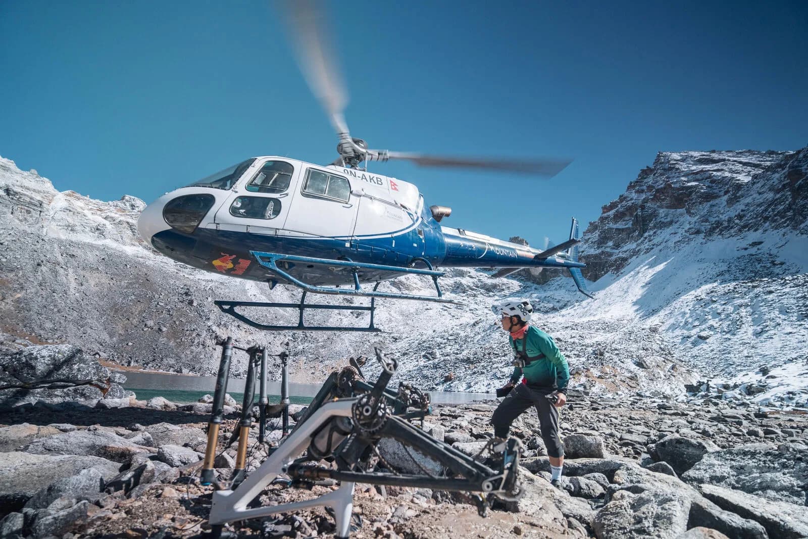 A helicopter comes in low over rocky, snow-dusted ground where bikes wait, a rider standing by.