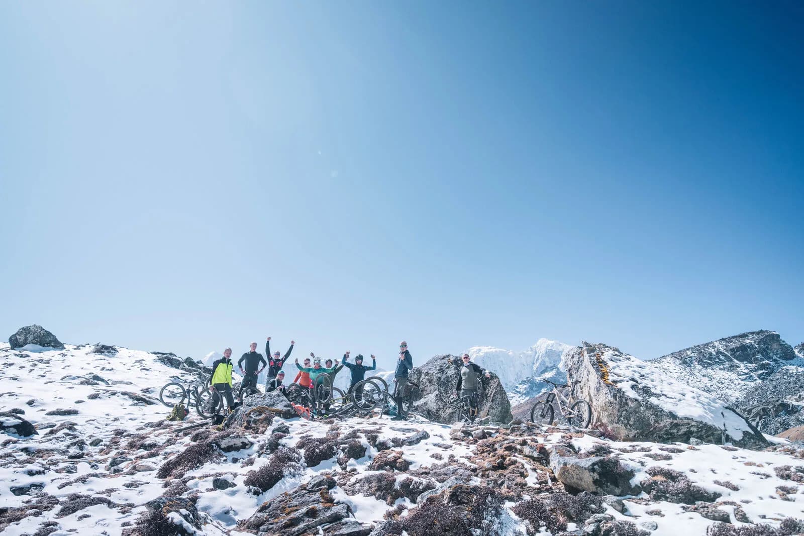 A group of riders raises their arms beside their bikes on snowy, rocky high ground.