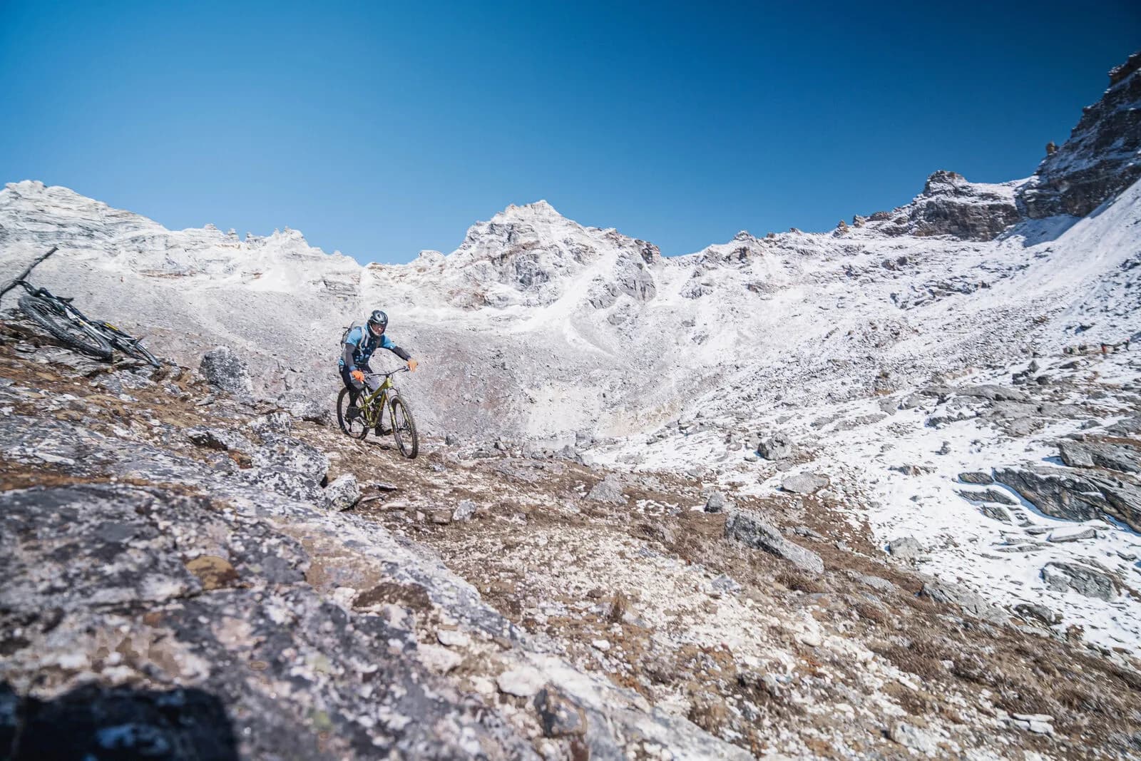 A rider picks a line down rocky, snow-dusted ground below white peaks.