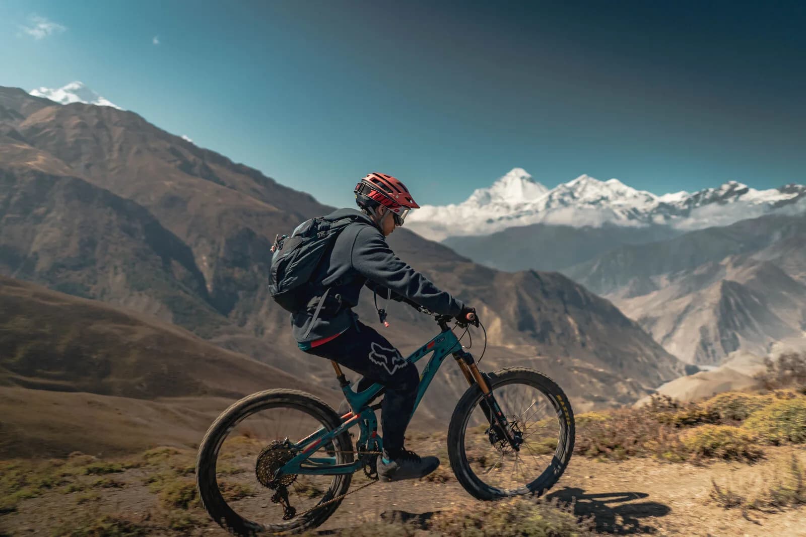 A rider in a red helmet rides across a high, dry slope, snow peaks on the horizon.