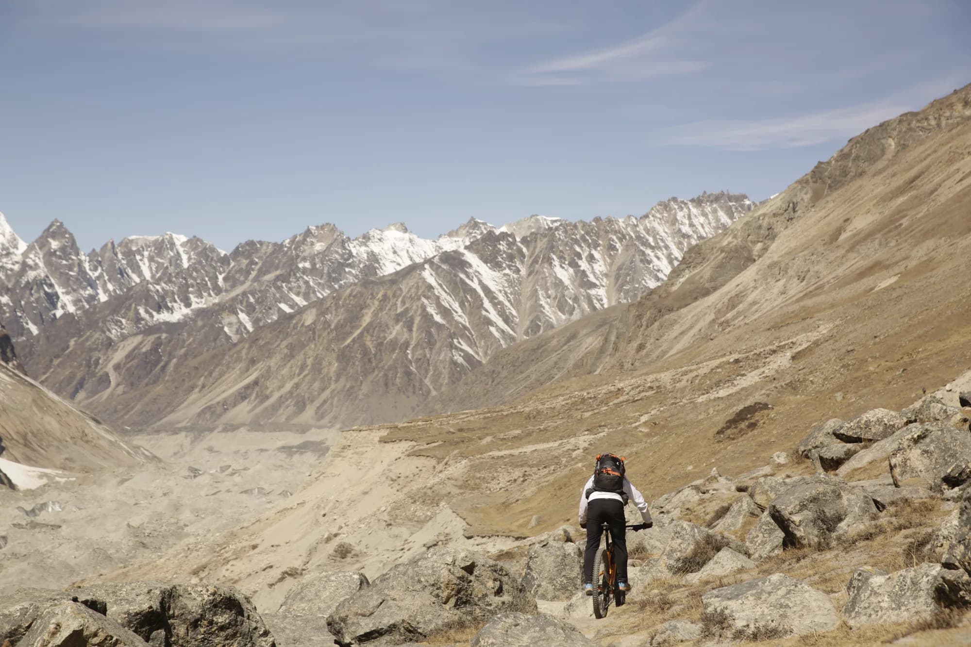 Mangal Krishna Lama riding a narrow stone trail high above a dry trans-Himalayan valley, with a long wall of snow-streaked peaks ahead.