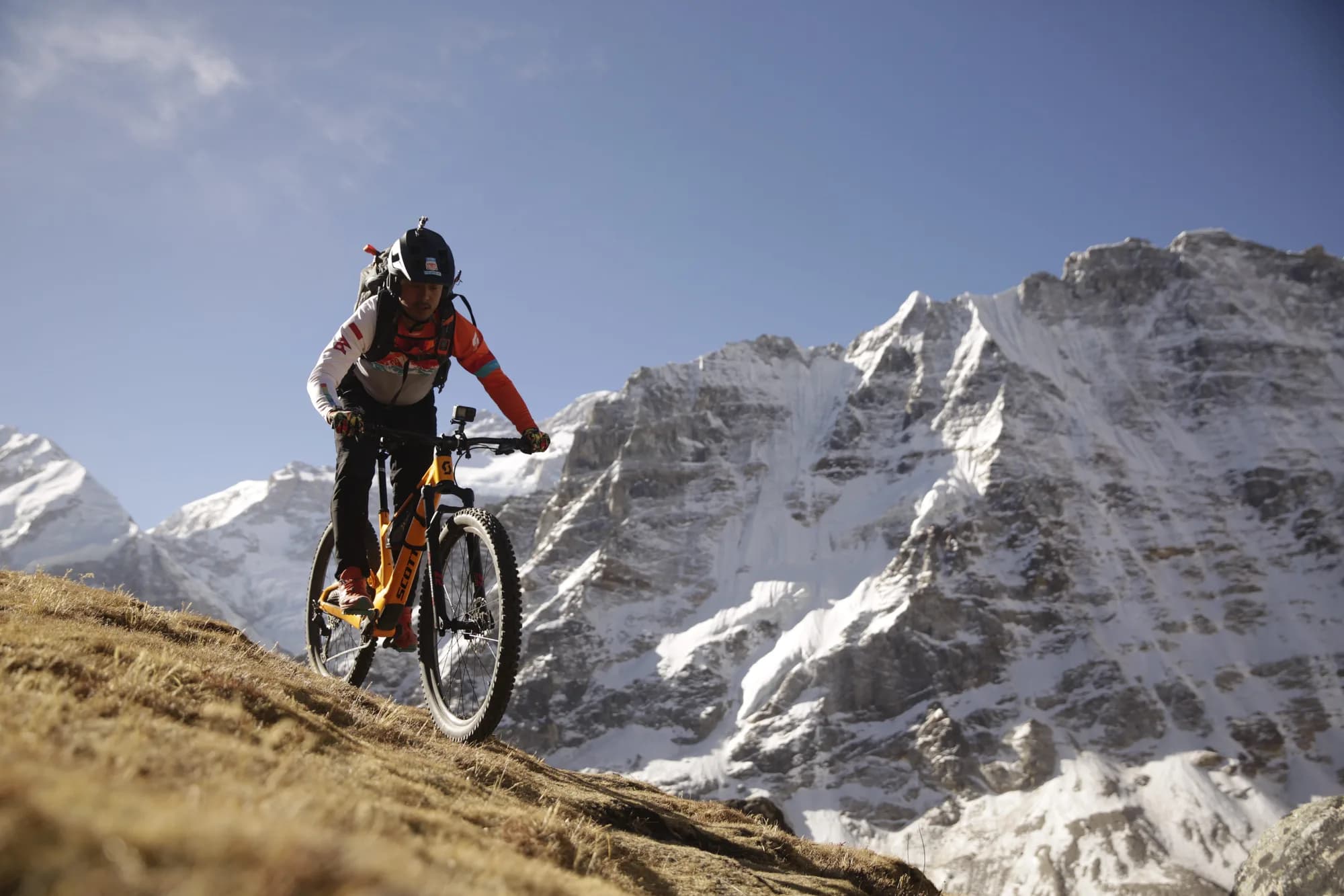 Mangal Krishna Lama riding his yellow full-suspension mountain bike across a steep golden alpine slope beneath a towering snow-covered Himalayan wall.