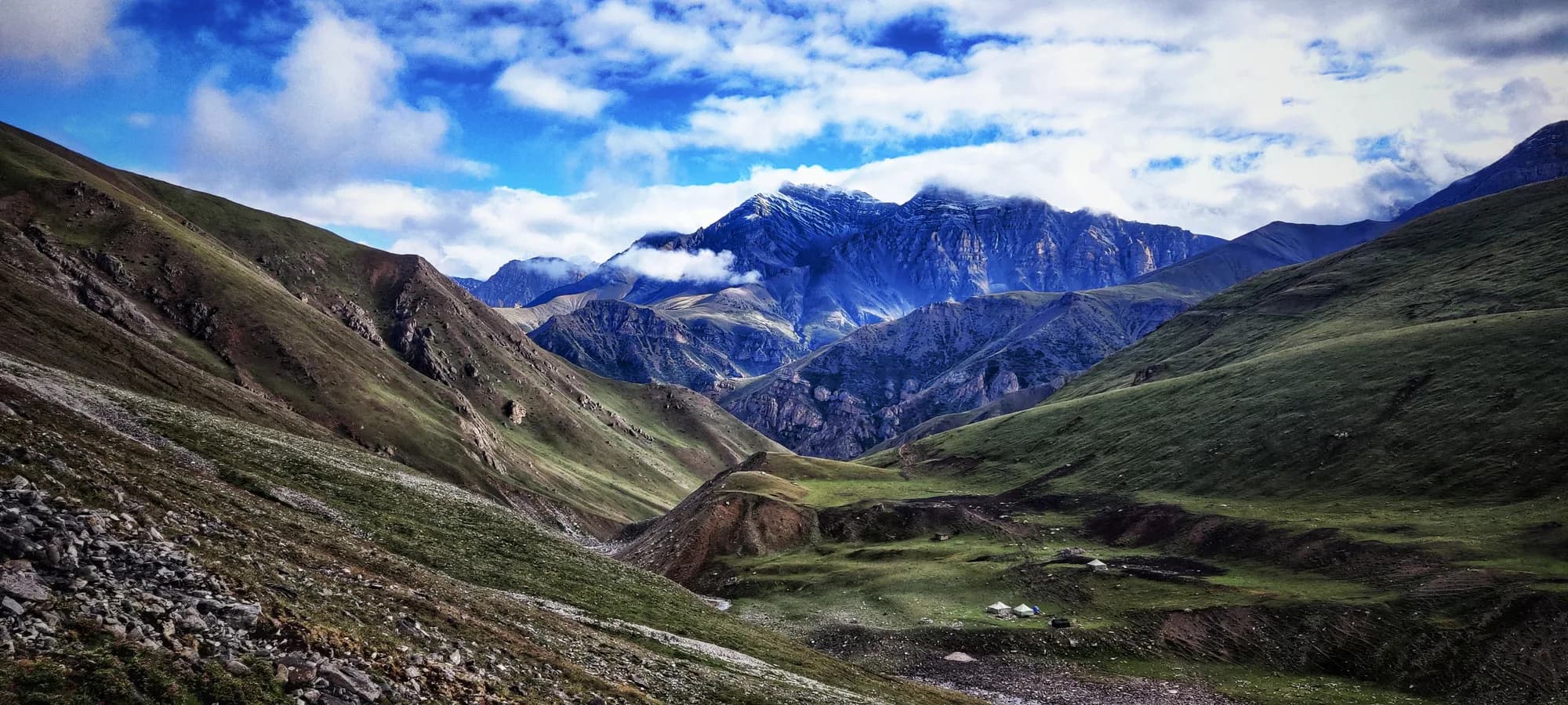 Nomad tents pitched in a green alpine valley floor between steep hillsides, with dark limestone peaks and clouds rising in the distance.