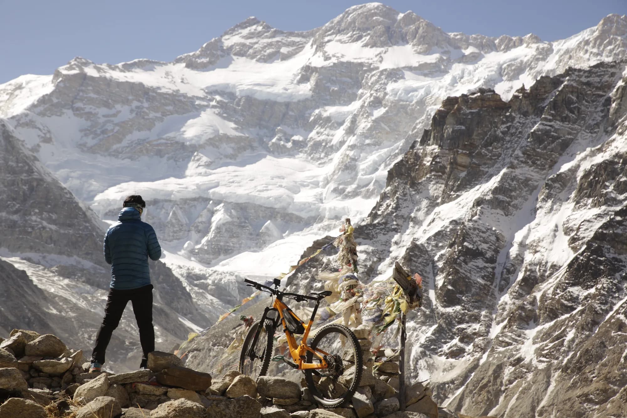 Mangal Krishna Lama standing beside his mountain bike and a prayer-flag chorten on a high Himalayan pass, with a glaciated snow wall rising behind him.