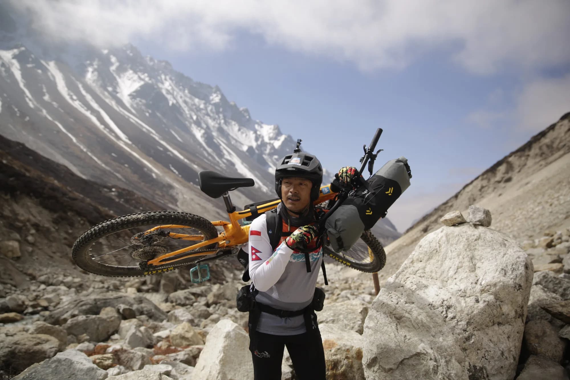 Mangal Krishna Lama carrying his mountain bike across his shoulders through a boulder-filled glacial moraine, with a dry-bag strapped to the handlebars and snow peaks behind.