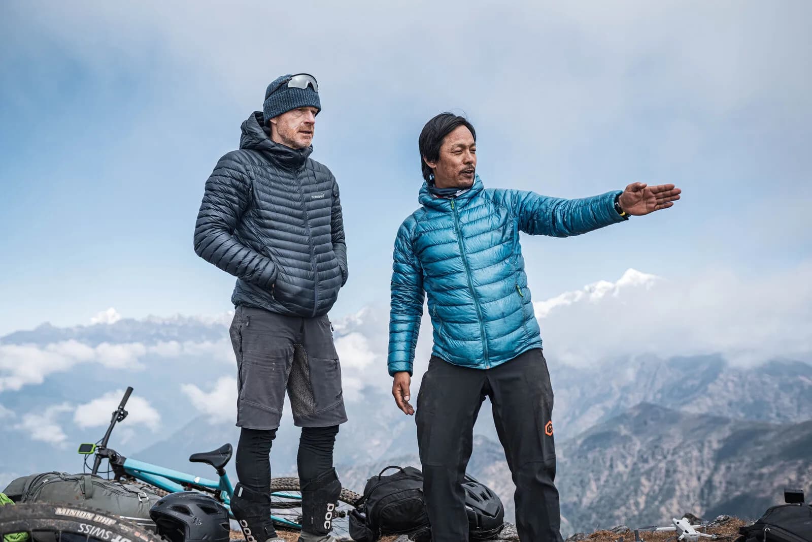 Mangal Lama, in a blue down jacket, points out a line across the range to a rider beside him; bikes and helmets lie on the ground behind.