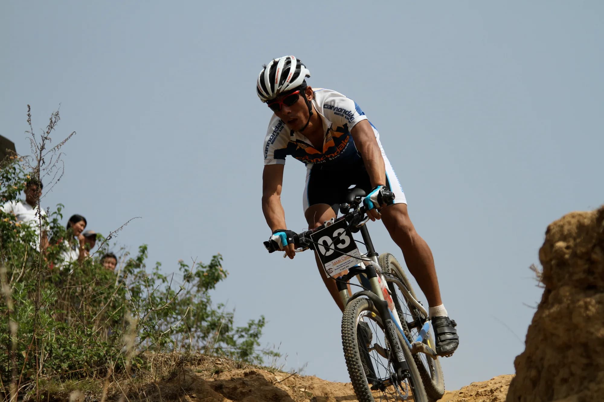 Mangal Lama racing a cross-country mountain bike over a dirt crest, a race number on his handlebars, spectators watching from the bank.