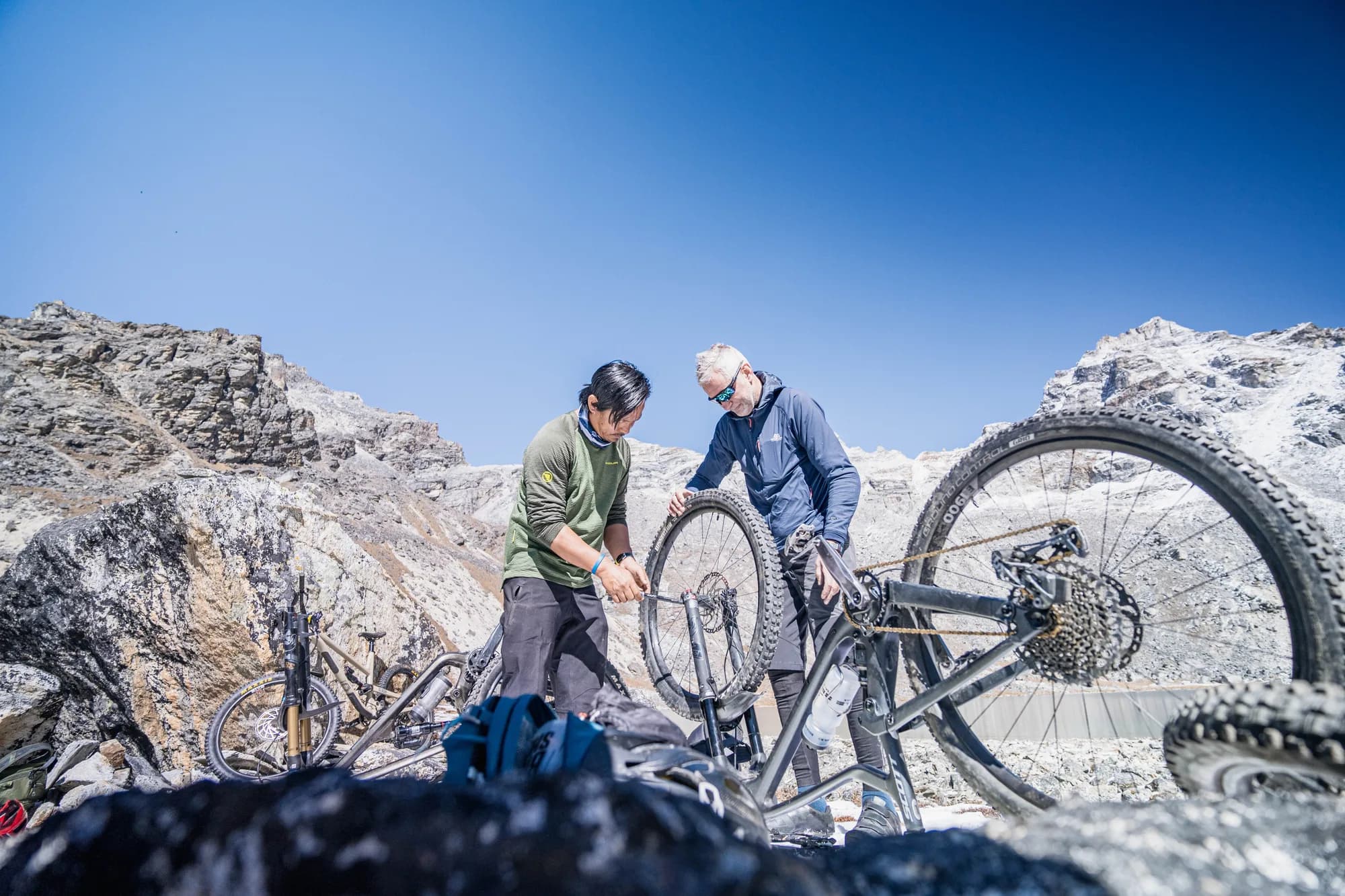 Mangal Lama works on a rider's bike among the boulders of a high valley, under a clear sky.