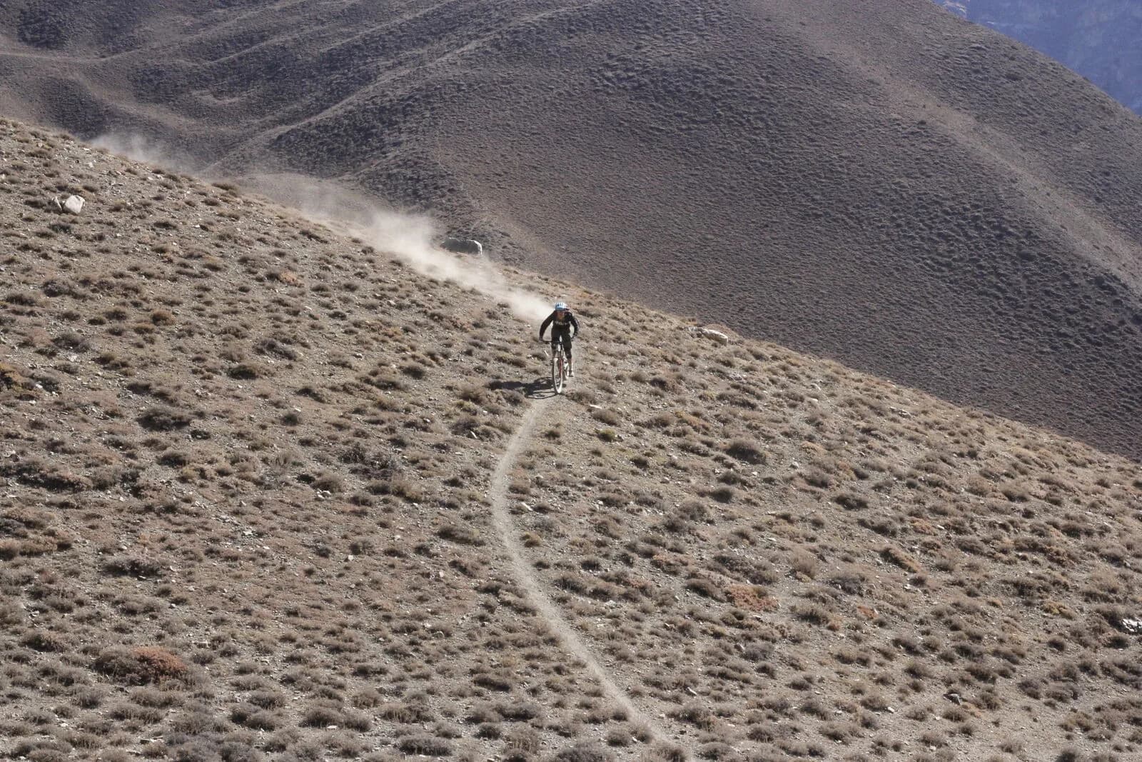 A lone mountain biker descends a thin singletrack across a vast dust-brown mountainside, a plume of dust rising behind.