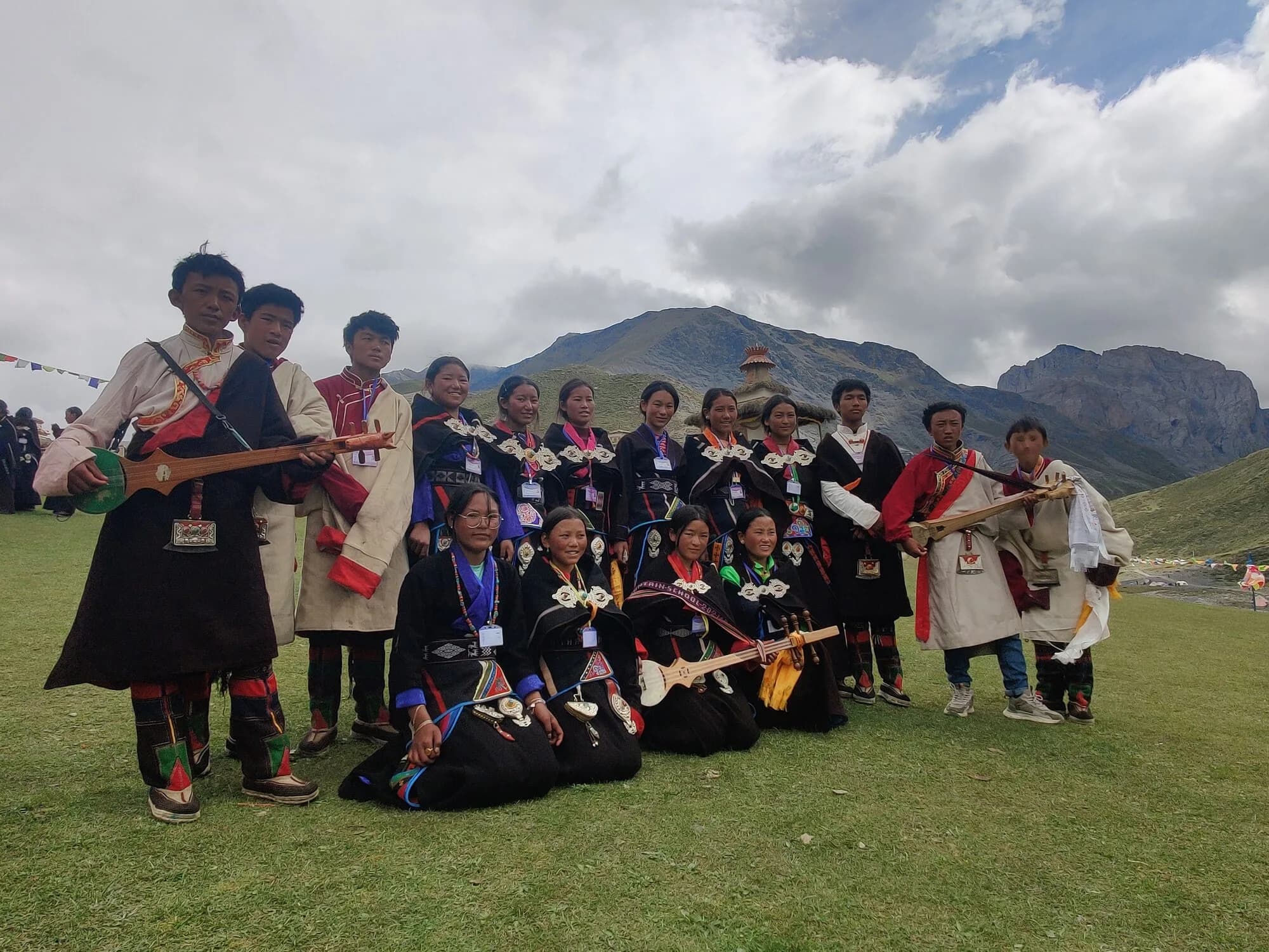 Young people in traditional dress with silver ornaments and lutes gather on festival grounds beneath grey peaks.
