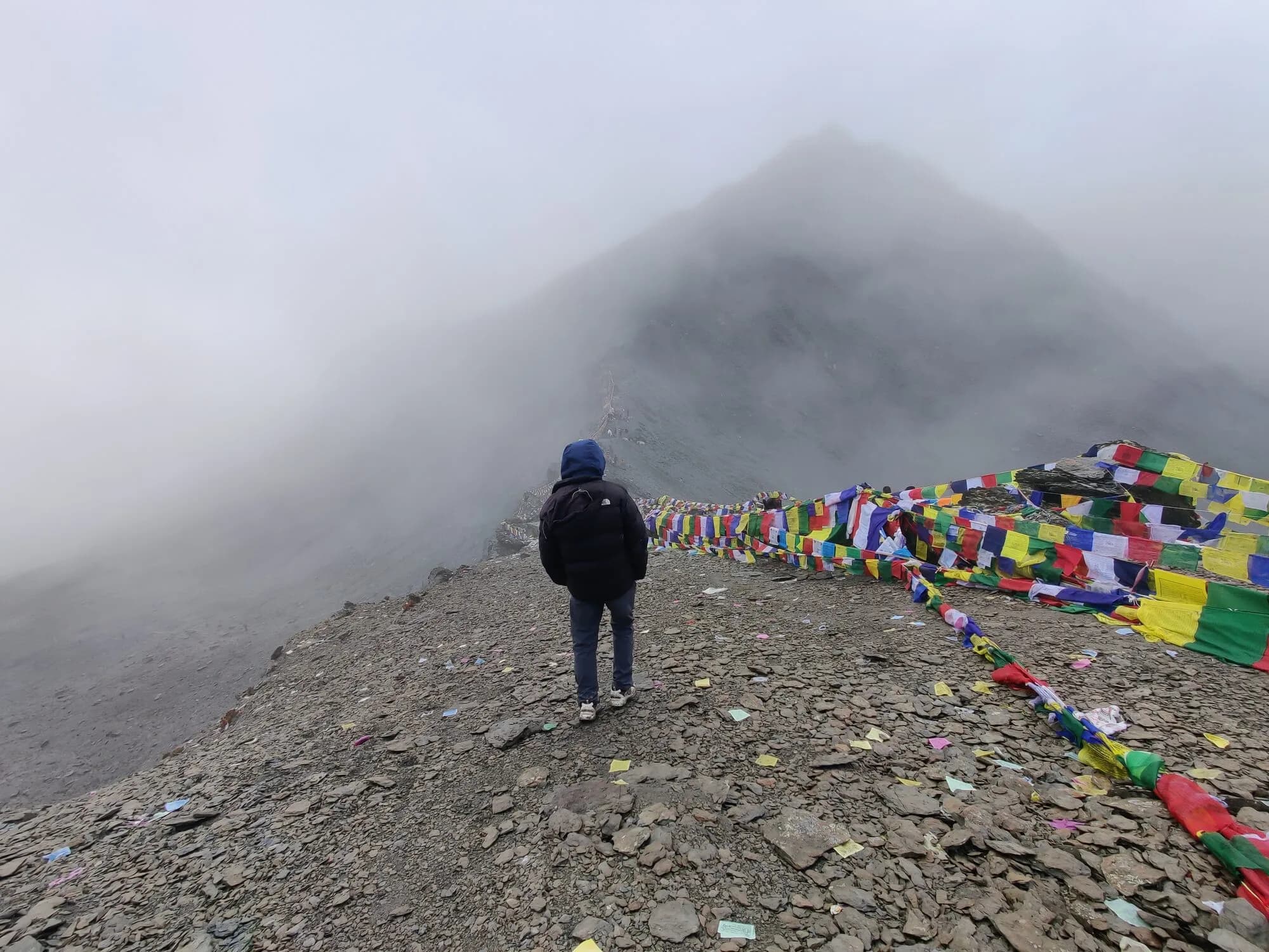 A hooded figure stands at a foggy scree pass beside masses of prayer flags, the ridge fading into cloud.