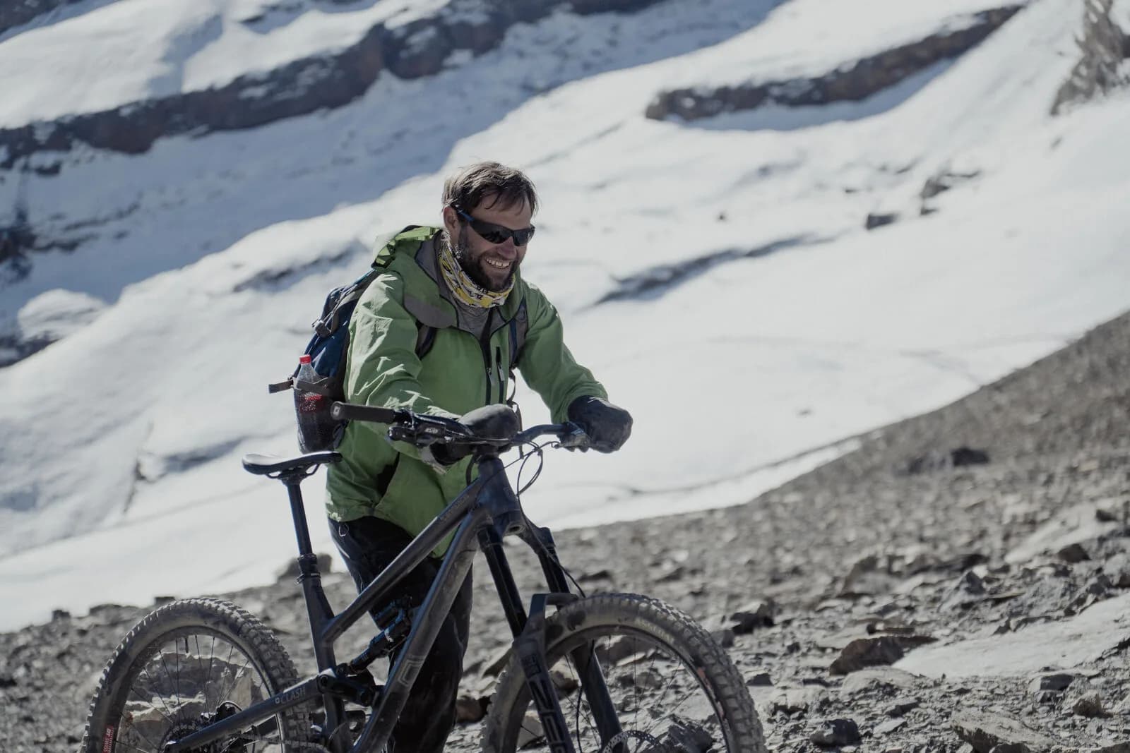 A smiling rider in a green jacket pushes a bike up scree below steep snowfields.