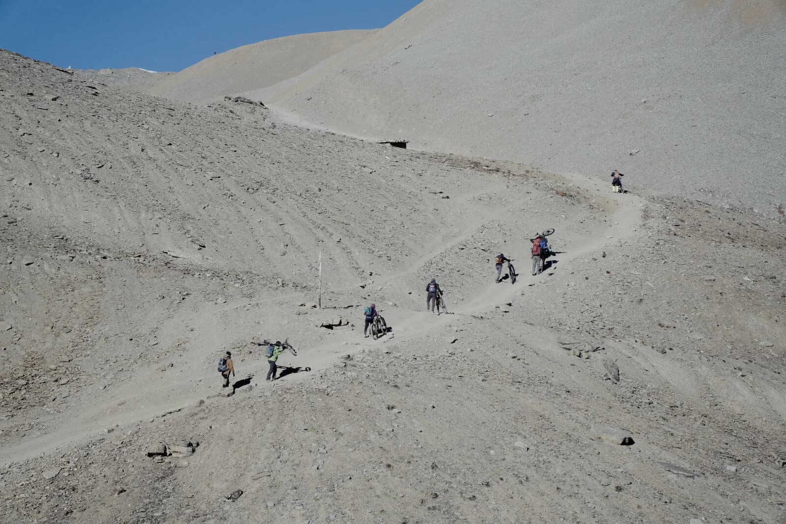A line of riders pushes and shoulders bikes up switchbacks cut into a bare scree mountainside.